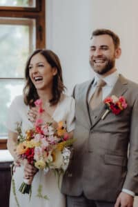 Ein fröhliches Paar steht drinnen zusammen und hält bunte Blumensträuße in den Händen. Die Frau trägt ein weißes Kleid und der Mann einen grauen Anzug mit beiger Krawatte. Beide lächeln breit, während sie ihre Hochzeit an einem Fenster feiern, durch das sanftes Tageslicht hereinströmt.
