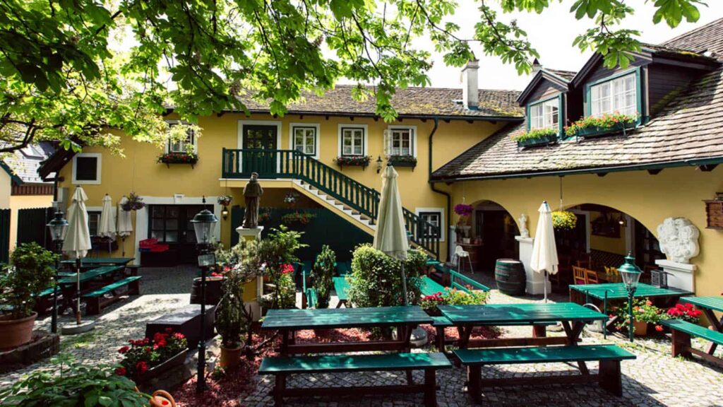 Charmanter Innenhof eines traditionellen Hauses im europäischen Stil mit gelber Fassade, perfekt für Hochzeitsfeiern. Genießen Sie grüne Bänke, bunte Blumen und Laubbäume. Mit Tischen und Sonnenschirmen im Freien und einer Treppe, die zu einem Balkon im zweiten Stock führt, der sich ideal für Hochzeitsfotos eignet.