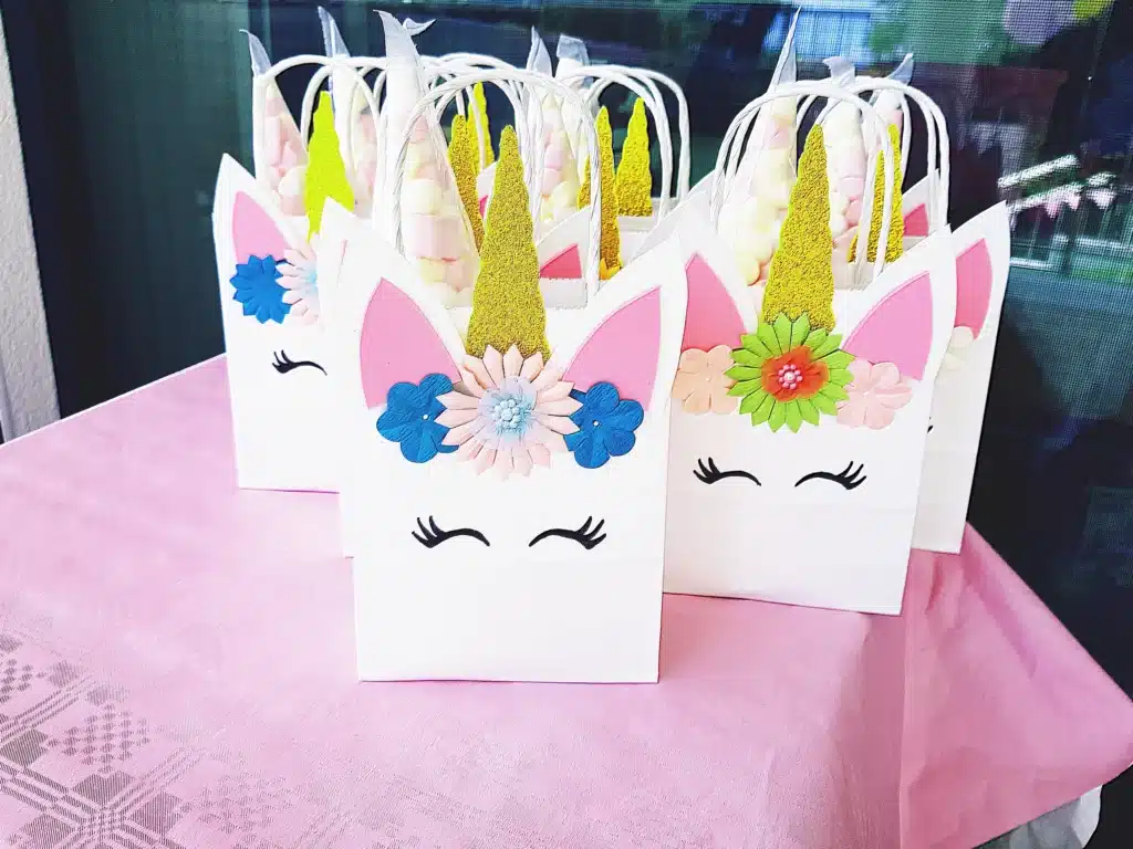 Eine Gruppe von Partygeschenktüten mit Einhornmotiven und bunten Papierohren, Hörnern und Blumen ist auf einer rosa Tischdecke arrangiert. Jede Tüte ist mit geschlossenen Augen und lebhaften Dekorationen versehen und eignet sich ideal, um jeder Hochzeitsfeier eine stimmungsvolle Note zu verleihen.