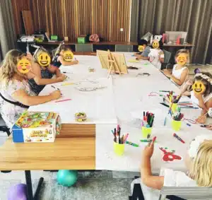 Eine Gruppe von Kindern sitzt um einen großen Tisch und beschäftigt sich mit Kunst- und Bastelaktivitäten, vielleicht um einer Hochzeitsfeier eine kreative Note zu verleihen. Der Tisch ist mit Papier und bunten Stiften bedeckt, an einem Ende steht eine Staffelei und festliche Luftballons liegen über den Boden verteilt.