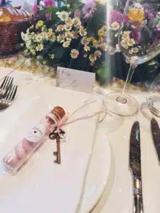 Ein perfekter Tisch für eine Hochzeit besteht aus einer Namenskarte, einem Weinglas, Besteck und einem Teller. Ein kleines Glas mit rosa Bonbons und ein mit einer Schleife zusammengebundener Schlüssel verleihen Charme. Im Hintergrund wertet ein Blumengesteck mit Gänseblümchen diese bezaubernde Szene auf, ideal für Ihre Hochzeitsplanung.