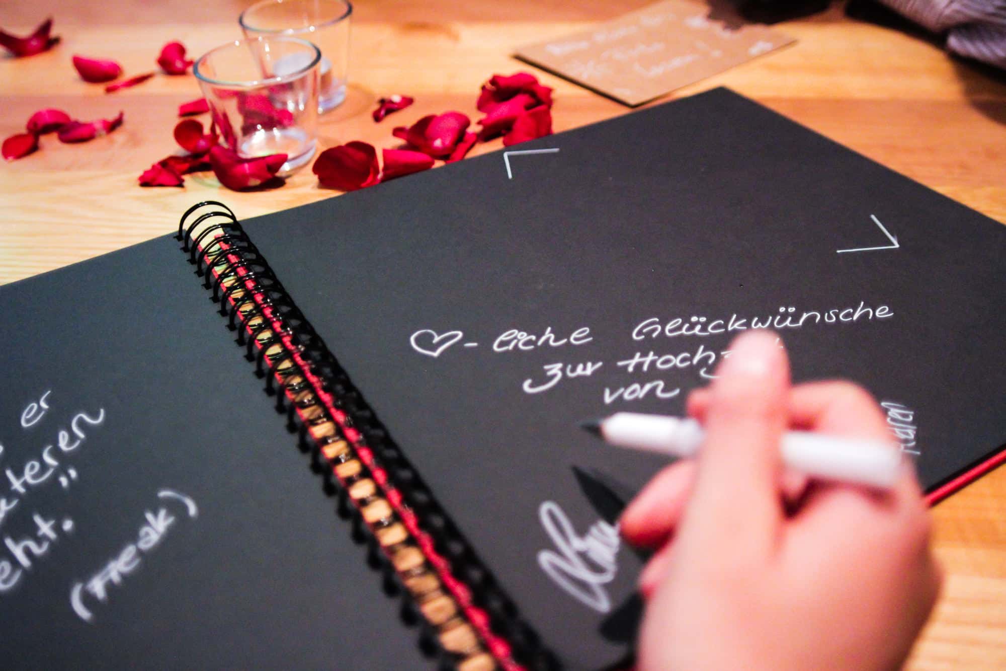 Eine Person schreibt mit einem weißen Stift in ein schwarzes Spiralnotizbuch. Die Seite enthält Glückwunschbotschaften auf Deutsch zur Feier einer Hochzeit. Rote Rosenblätter und kleine Kerzen sind auf dem Holztisch rund um das Notizbuch verstreut und schaffen eine romantische Atmosphäre für die Planung des perfekten Anlasses.