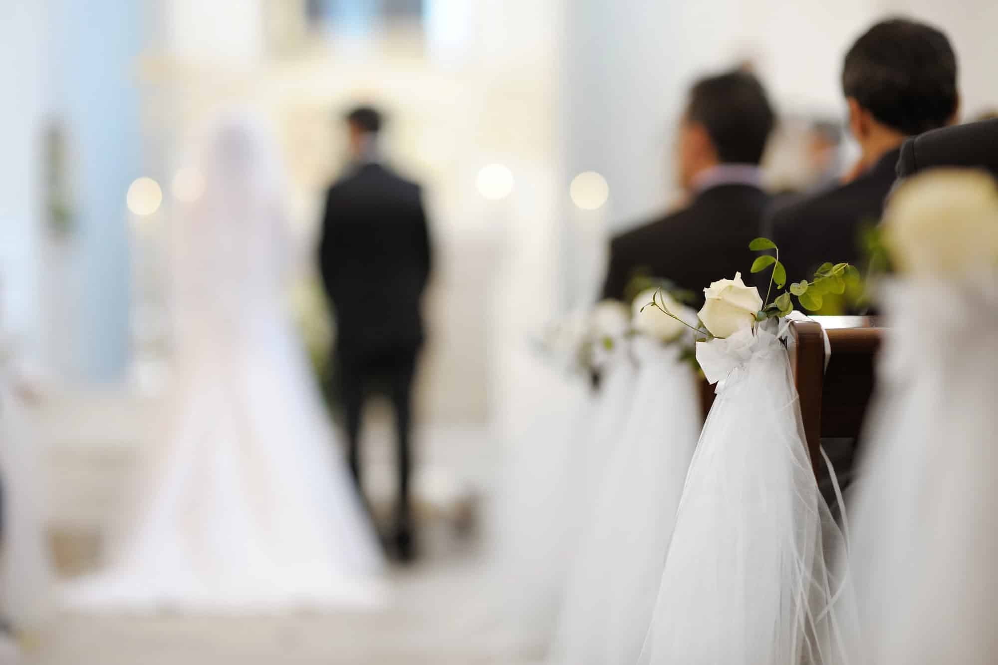 Eine verschwommene Ansicht einer Hochzeitszeremonie zeigt eine Braut in einem weißen Kleid und einen Bräutigam in einem schwarzen Anzug am Altar. Weiße Rosen und Tülldekorationen schmücken die Kirchenbänke im Vordergrund und schaffen eine bezaubernde Atmosphäre für diesen besonderen Hochzeitsmoment.