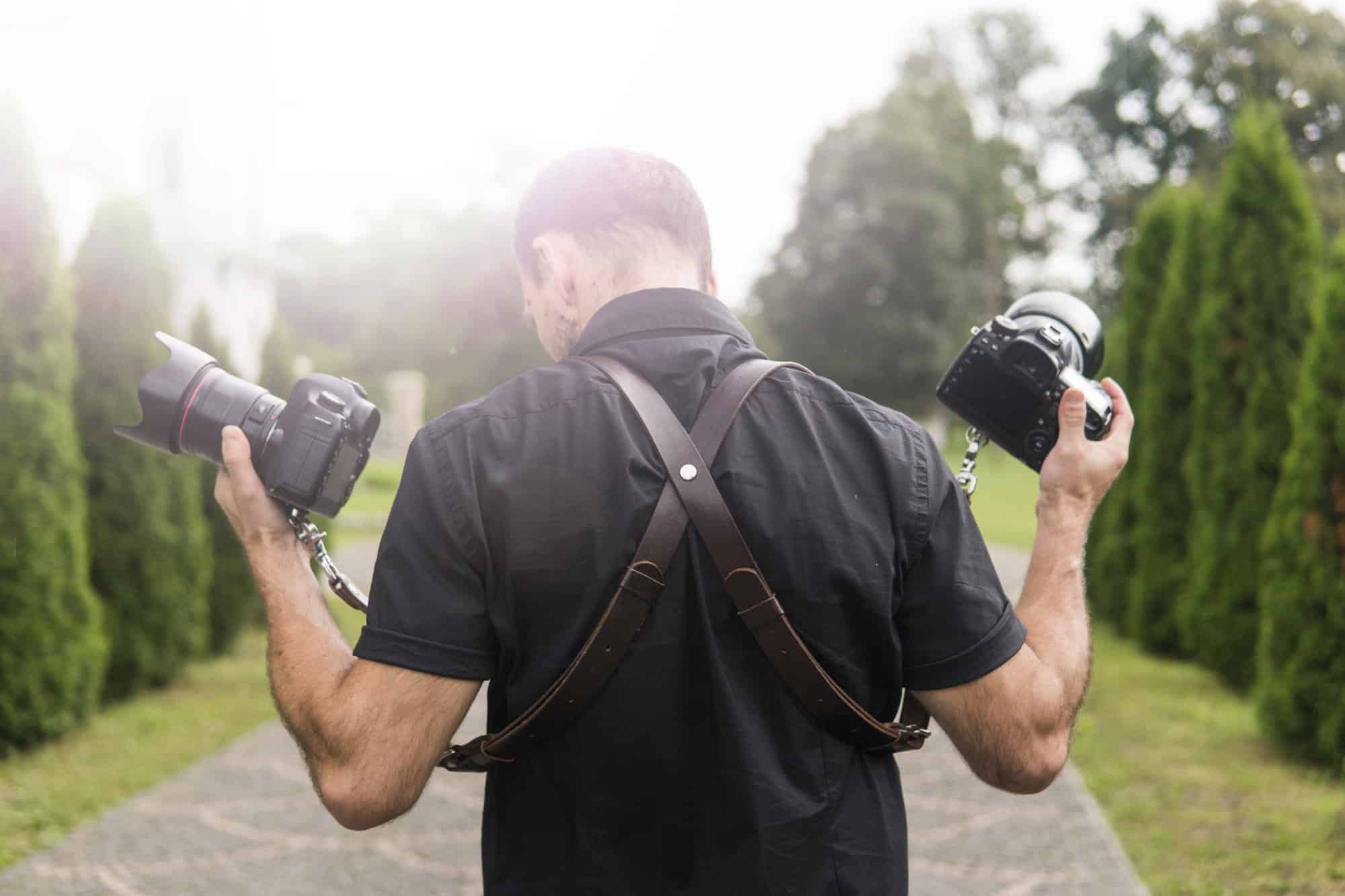 Eine Person in einem schwarzen Hemd steht draußen auf einem Weg und hält zwei Kameras mit einem Ledergeschirr über den Schultern. Hohe Bäume und viel Grün säumen den Weg und schaffen eine perfekte Kulisse, um bezaubernde Momente von Paaren festzuhalten, die ihre Hochzeit inmitten des strahlenden Tageslichts planen.