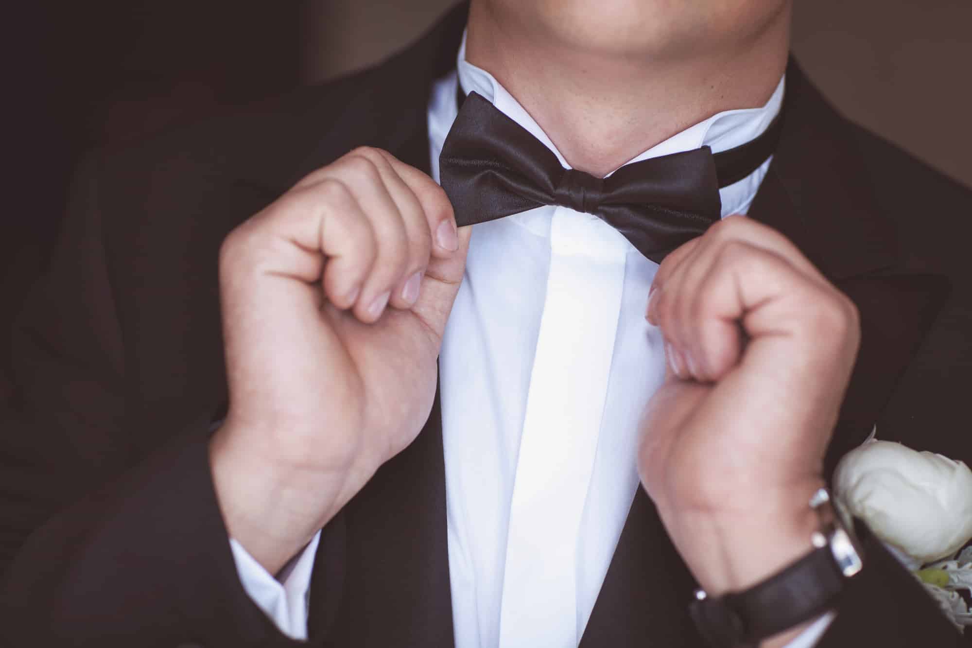 Eine Person, die einen dunklen Anzug trägt, richtet eine schwarze Fliege an einem weißen Oberhemd. Am Revers der Jacke ist eine weiße Ansteckblume befestigt, die die Eleganz der Hochzeitsvorbereitungen verkörpert.