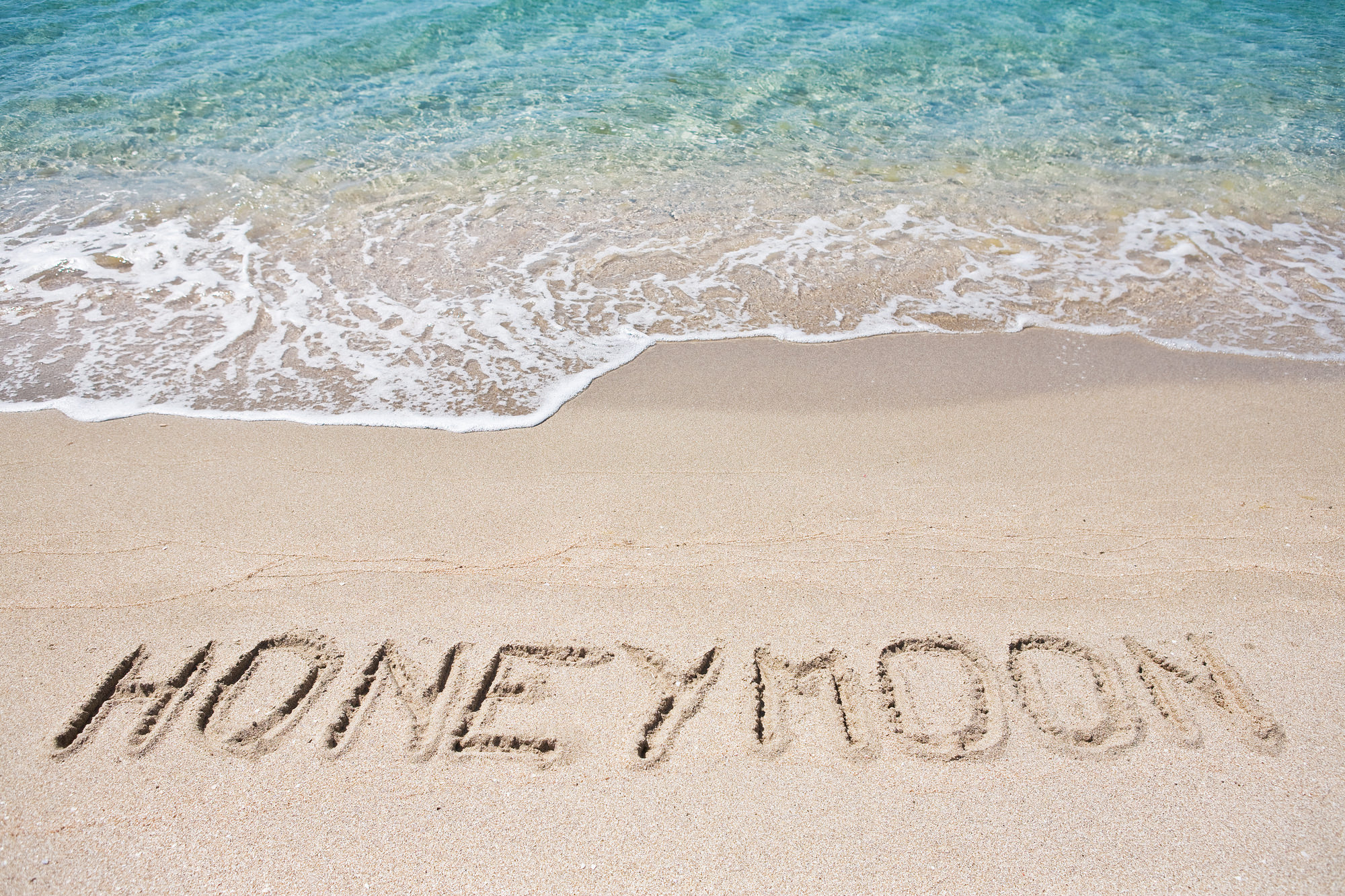 Das Wort „Flitterwochen“ ist in den Sand eines Strandes geschrieben, an dem die Wellen sanft ans Ufer heranrollen. Das leuchtende Blau des Ozeans unterstreicht diese ruhige Szene, perfekt für ein Paar, das gerade erst in die Hochzeitsplanung eingetaucht ist und seine ersten Momente des Eheglücks genießt.