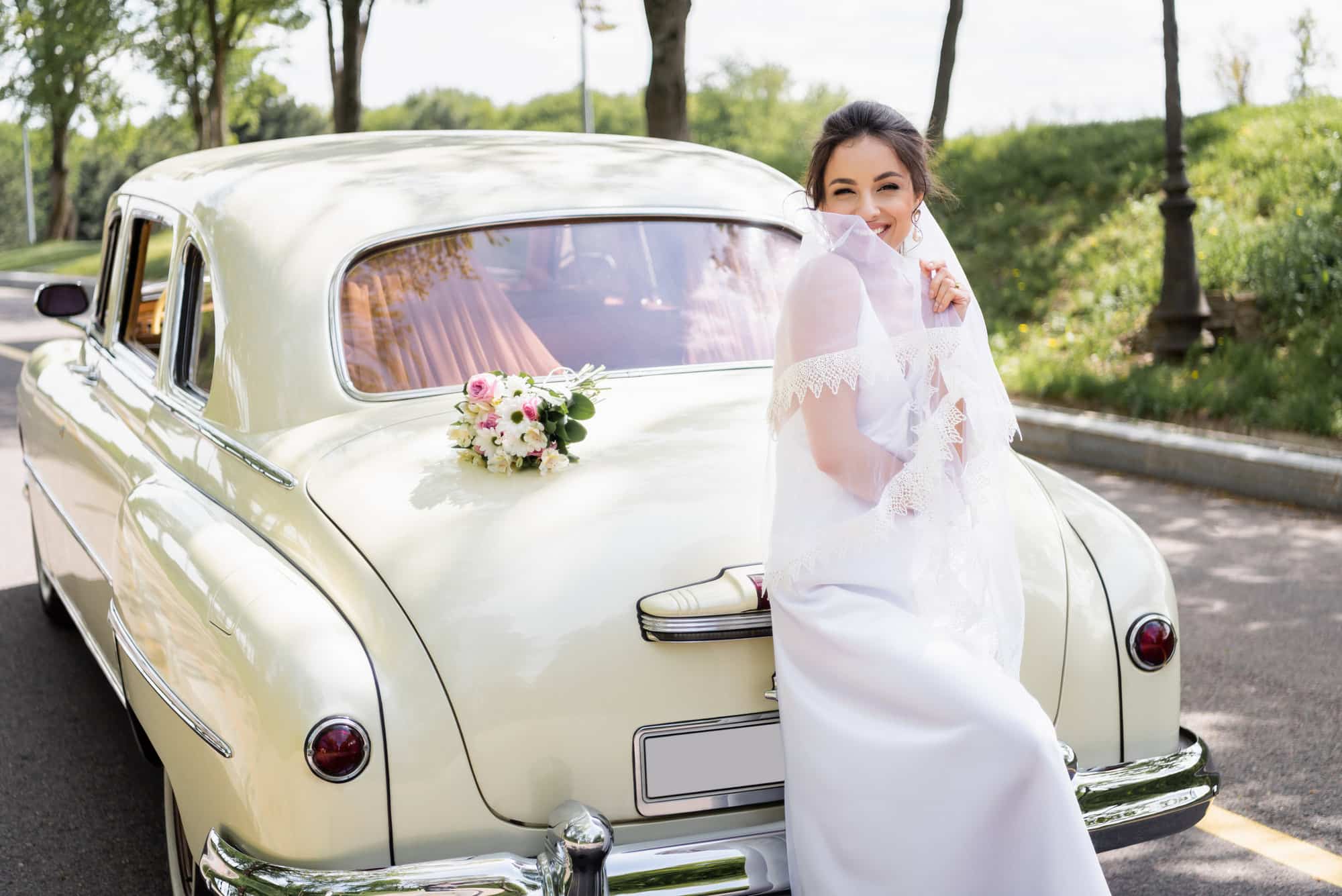 Eine Braut in einem weißen Kleid und Schleier sitzt auf dem Kofferraum eines alten Hochzeitsautos, hält einen Teil ihres Schleiers und lächelt. Ein Strauß weißer und rosa Blumen liegt auf dem Auto, während im Hintergrund Bäume die Straße säumen.
