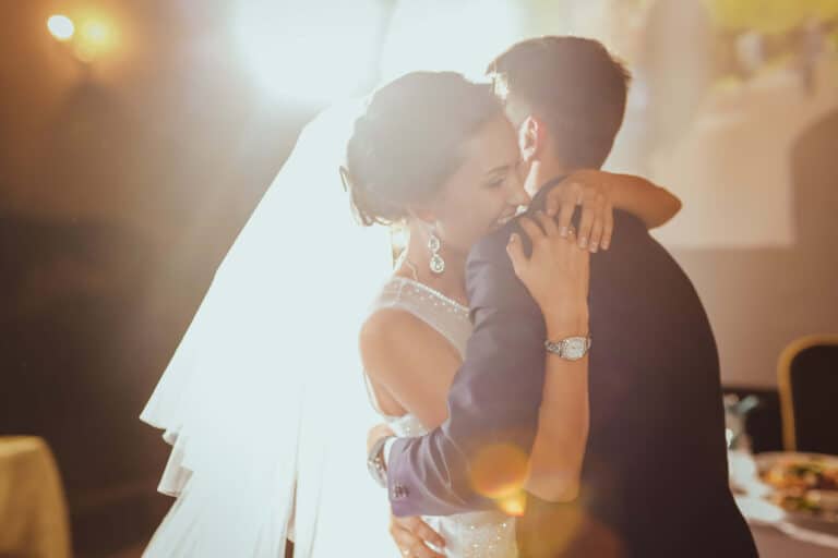 Eine Braut in weißem Kleid und Schleier umarmt einen Bräutigam im dunklen Anzug. Sie tanzen eng aneinander gekuschelt in einem warm beleuchteten Raum und verkörpern die Freude ihrer Hochzeit. Lichtreflexe verleihen dem Bild einen verträumten Effekt und betonen die Intimität und Feierlichkeit des Augenblicks der Hochzeit.