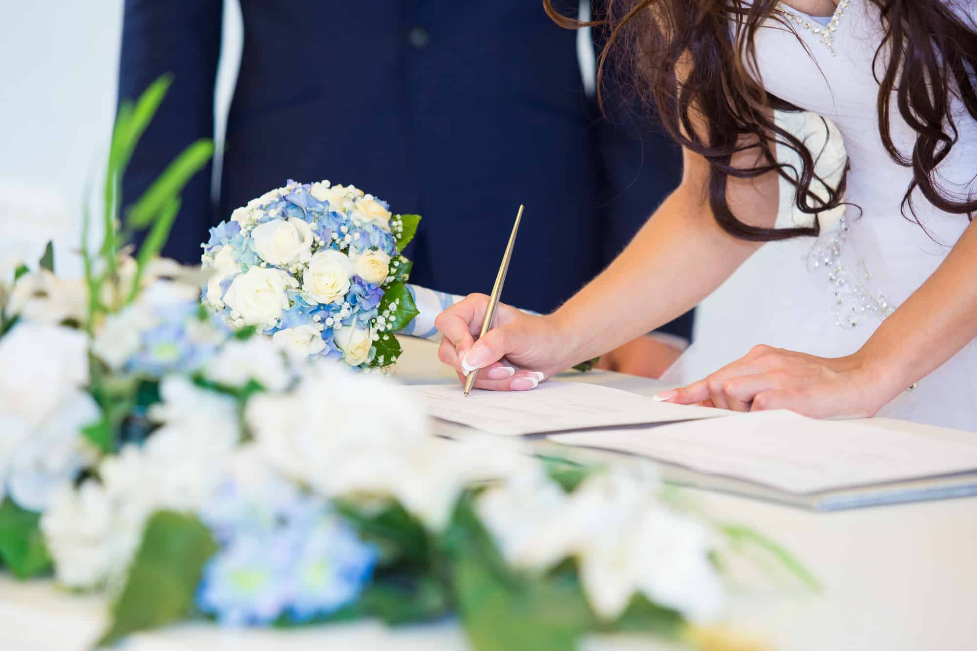 Eine Braut unterschreibt ein Dokument bei ihrer Hochzeit und hält dabei einen Stift in ihrer rechten Hand. Der Fokus liegt auf ihrer Hand und dem Strauß blauer und weißer Blumen auf dem Tisch. Der Bräutigam steht neben ihr und trägt einen dunklen Anzug, was ihre sorgfältige Hochzeitsplanung widerspiegelt.