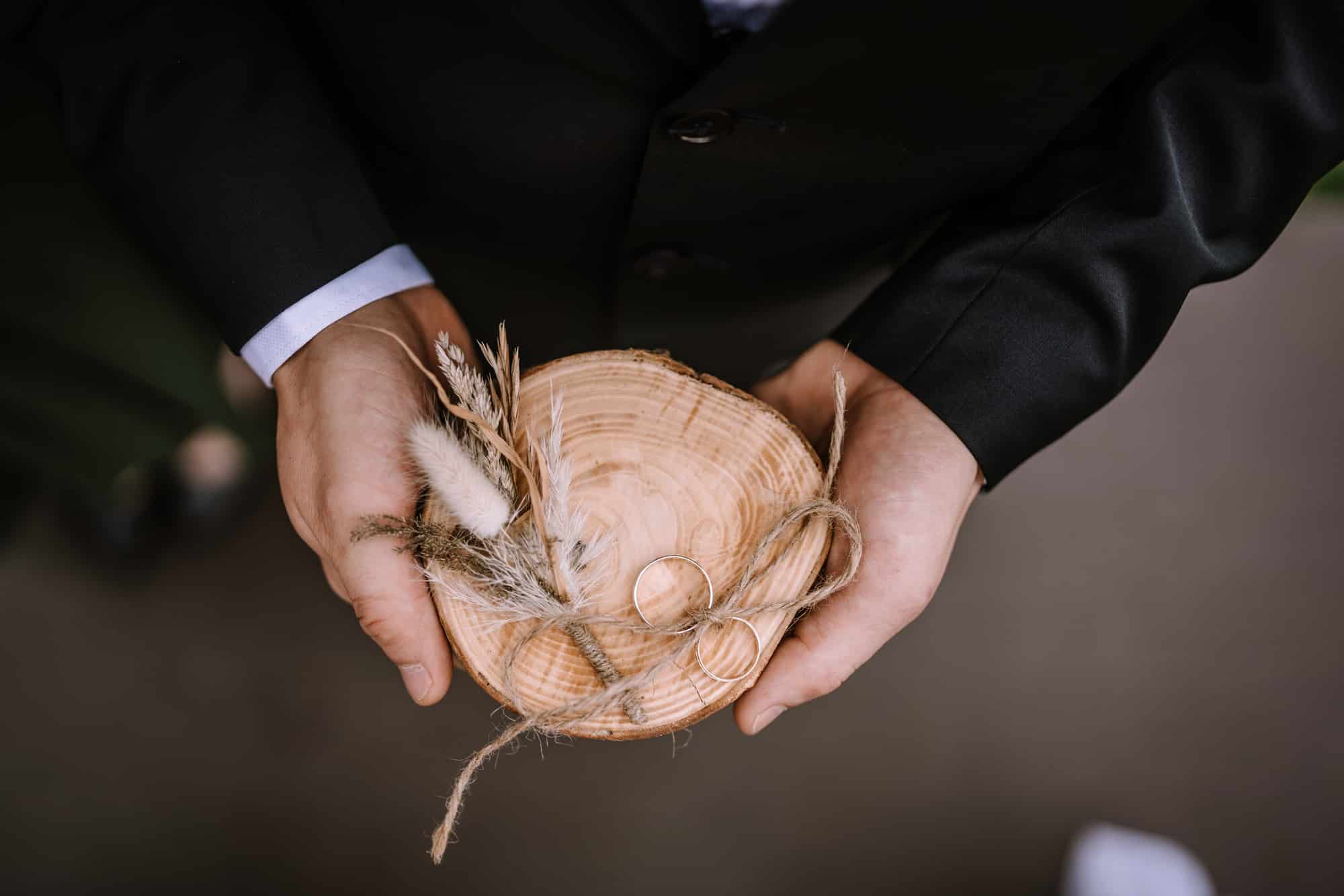 Eine Person im schwarzen Anzug hält eine Holzplatte mit zwei Eheringen, die mit rustikaler Schnur zusammengebunden sind und die Essenz der Hochzeitsplanung verkörpern. Ein kleiner Zweig getrockneter Blumen ist vorsichtig neben den Ringen auf der Platte platziert.