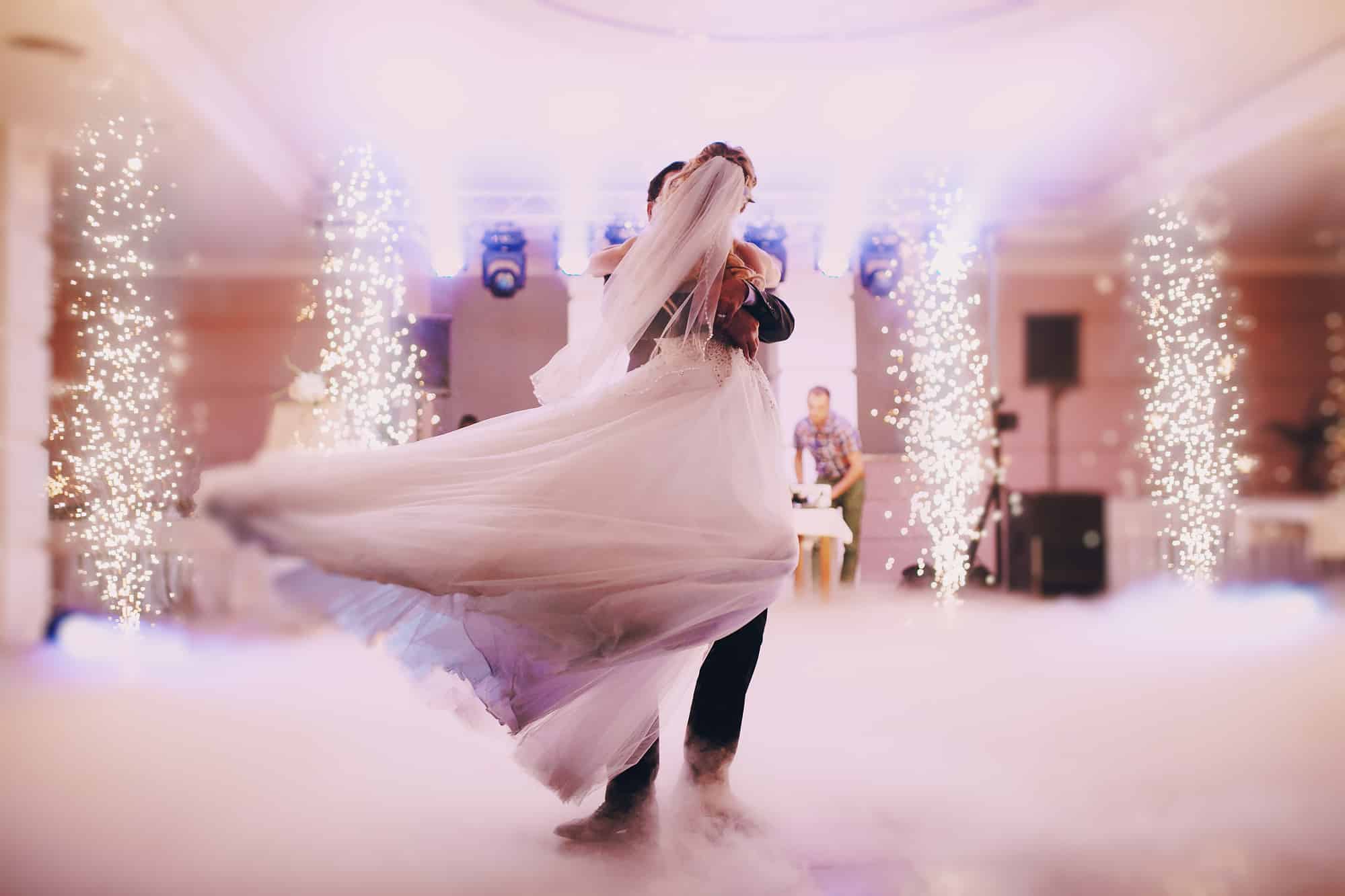 Ein frisch vermähltes Paar genießt seinen Hochzeitstanz in einer traumhaften Umgebung, umgeben von Nebel und funkelnden Lichtern. Das fließende Kleid der Braut wird mitten in der Drehung eingefangen und schafft eine bezaubernde Atmosphäre. Der Hintergrund ist sanft beleuchtet und trägt zur magischen Szene bei.
