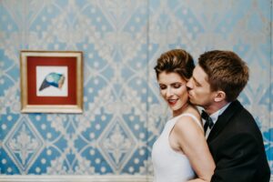 Ein Paar steht eng aneinander gedrängt vor einer gemusterten blauen Wand und verkörpert die Essenz einer Hochzeit. Der Mann im Anzug umarmt die Frau im ärmellosen weißen Kleid und küsst sie sanft auf die Wange. Neben ihnen hängt ein gerahmtes Bild eines Vogels, das diesem intimen Moment noch mehr Charme verleiht.