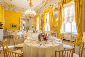 Der luxuriöse Speisesaal, der sich perfekt für eine Hochzeit eignet, besticht durch elegante Kronleuchter und goldenes Dekor. Runde Tische sind mit feinem Porzellan und Blumendekorationen gedeckt und von goldenen Stühlen umgeben. Große Fenster mit drapierten Vorhängen lassen natürliches Licht herein und schaffen eine opulente Atmosphäre, die ideal für die Hochzeitsplanung ist.