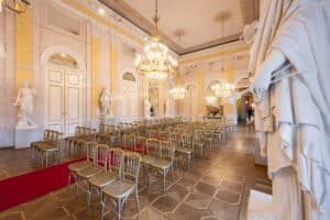 Ein eleganter Ballsaal mit kunstvollen Kronleuchtern, goldenen Stühlen in Reihen und einem Gang mit rotem Teppich bietet den perfekten Rahmen für eine Hochzeit. Große Statuen und kunstvolle Zierleisten schmücken die Wände, während hohe Fenster die großartige, historische Atmosphäre verstärken, die ideal für die Hochzeitsplanung ist.