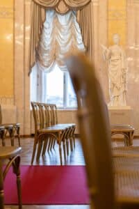 Ein eleganter Raum bietet die perfekte Kulisse für eine Hochzeit, mit drapierten Vorhängen, leeren Stühlen und einer Statue im Hintergrund. Der Boden ist mit einem roten Teppich geschmückt, und durch ein Fenster fällt natürliches Licht herein – ideal für die Hochzeitsplanung.