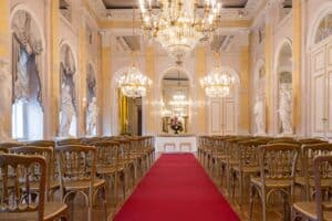 Ein großer Saal mit eleganten Kronleuchtern und einem Gang mit rotem Teppich, flankiert von Reihen verzierter goldener Stühle. Statuen und Spiegel schmücken die Wände und schaffen eine luxuriöse Atmosphäre, die perfekt für eine Hochzeit oder jede formelle Veranstaltung ist.
