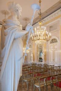 Ein großer Saal mit Kronleuchtern und kunstvollem Dekor verfügt über Reihen goldener Stühle mit roten Kissen, perfekt für eine Traumhochzeit. Eine große Marmorstatue mit einem Musikinstrument steht im Vordergrund und verleiht der opulenten Kulisse eine elegante Note, ideal für anspruchsvolle Hochzeitsplanung.