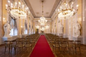 Ein großer Saal mit einem roten Teppich als Mittelgang, perfekt für eine Hochzeit, flankiert von Reihen verzierter Stühle. Der Raum verfügt über hohe Fenster, große Kronleuchter und Statuen an den Wänden, die eine elegante, klassische Atmosphäre schaffen, ideal für anspruchsvolle Hochzeitsplanung.