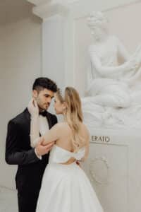 Ein Paar in formeller Kleidung genießt einen romantischen Moment vor einer klassischen Statue, perfekt für ihre Hochzeit. Der Mann trägt einen schwarzen Smoking und die Frau ein weißes Kleid mit Schleife. Die Statue mit dem Namen „ERATO“ hält ein Musikinstrument und verleiht ihrer Hochzeitsszene Harmonie.
