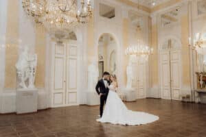 Braut und Bräutigam stehen in einem eleganten Ballsaal, der mit Marmorstatuen, Kronleuchtern und hohen Decken geschmückt ist und ihre Hochzeitsträume verkörpert. Die Braut trägt ein weißes Kleid, der Bräutigam einen schwarzen Anzug. Sie umarmen sich herzlich in der Mitte dieser exquisiten Kulisse, perfekt für ihren Hochzeitsmoment.