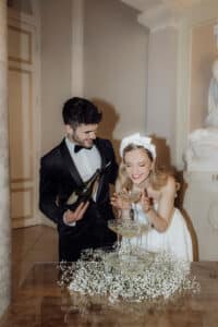 Das Paar feiert seine Hochzeit bei einer formellen Veranstaltung. Der Mann im Smoking gießt Champagner in einen Turm aus Gläsern, während die Frau im weißen Kleid und mit Haarreif freudig die Hände ausstreckt. Die Szene, perfekt für Inspiration bei der Hochzeitsplanung, ist mit weißen Blumen geschmückt.