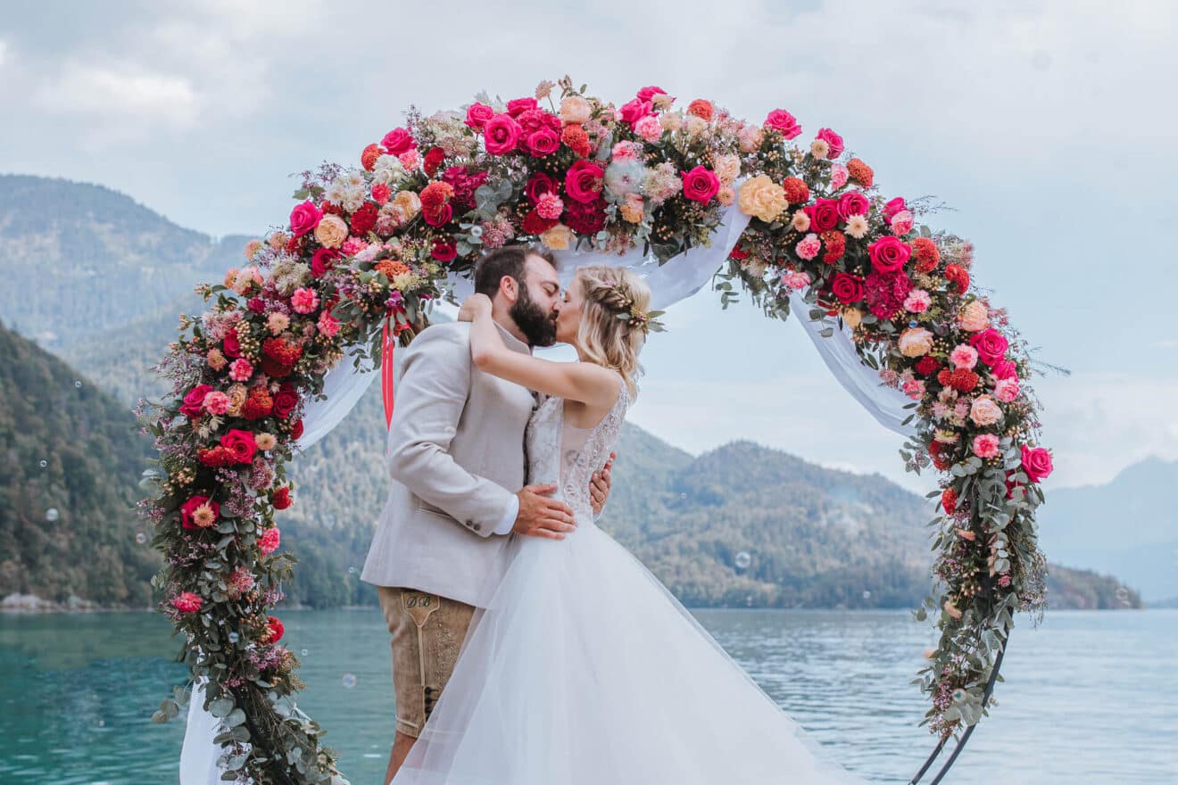 Braut und Bräutigam küssen sich unter einem mit roten und rosa Rosen geschmückten Blumenbogen an einem ruhigen, von Bergen umgebenen See – ein malerischer Moment, perfekt zum Heiraten. Die Braut trägt ein weißes Kleid und der Bräutigam einen beigen Anzug. Der Himmel ist bewölkt, aber hell und bietet die ideale Kulisse für ihre Hochzeit.