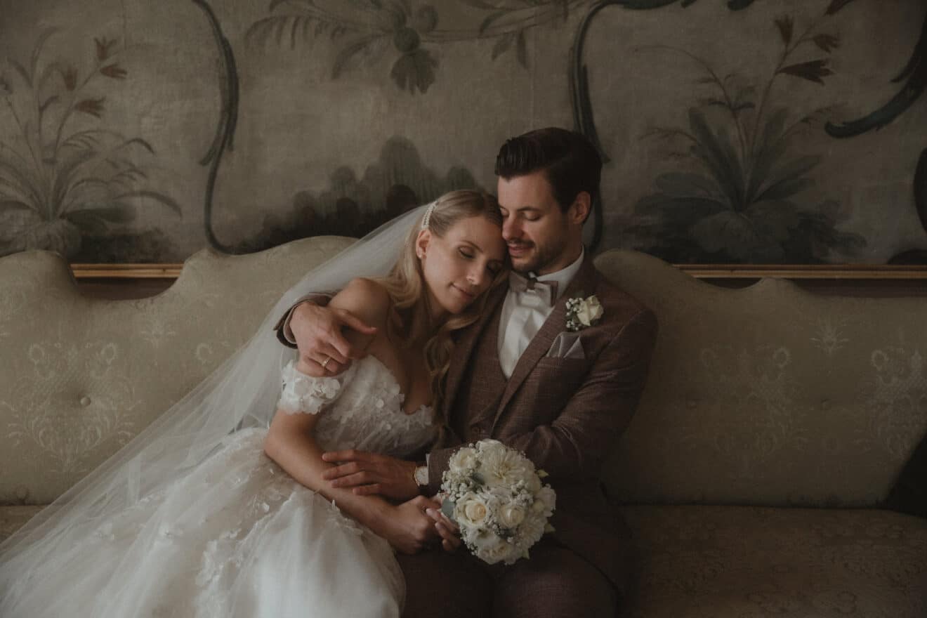 Braut und Bräutigam sitzen eng beieinander auf einem eleganten Sofa und sind in ihre Hochzeitsplanung vertieft. Die Braut, in einem weißen Kleid und mit einem Blumenstrauß in der Hand, legt ihren Kopf auf die Schulter des Bräutigams. Er, in einem braunen Anzug, umarmt sie vor der Kulisse einer reich verzierten Blumentapete.