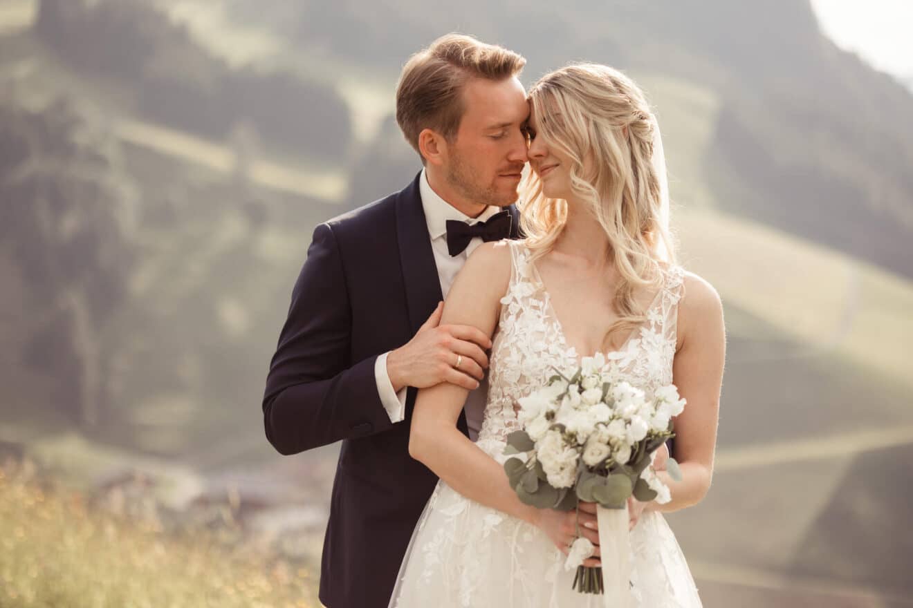 Braut und Bräutigam umarmen sich in einer malerischen Umgebung im Freien und feiern ihre Hochzeit. Der Bräutigam trägt einen dunklen Anzug und eine Fliege, während die Braut ein weißes Spitzenkleid trägt und einen Strauß weißer Blumen hält. Im Hintergrund sind verschwommene, sanfte Hügel zu sehen.