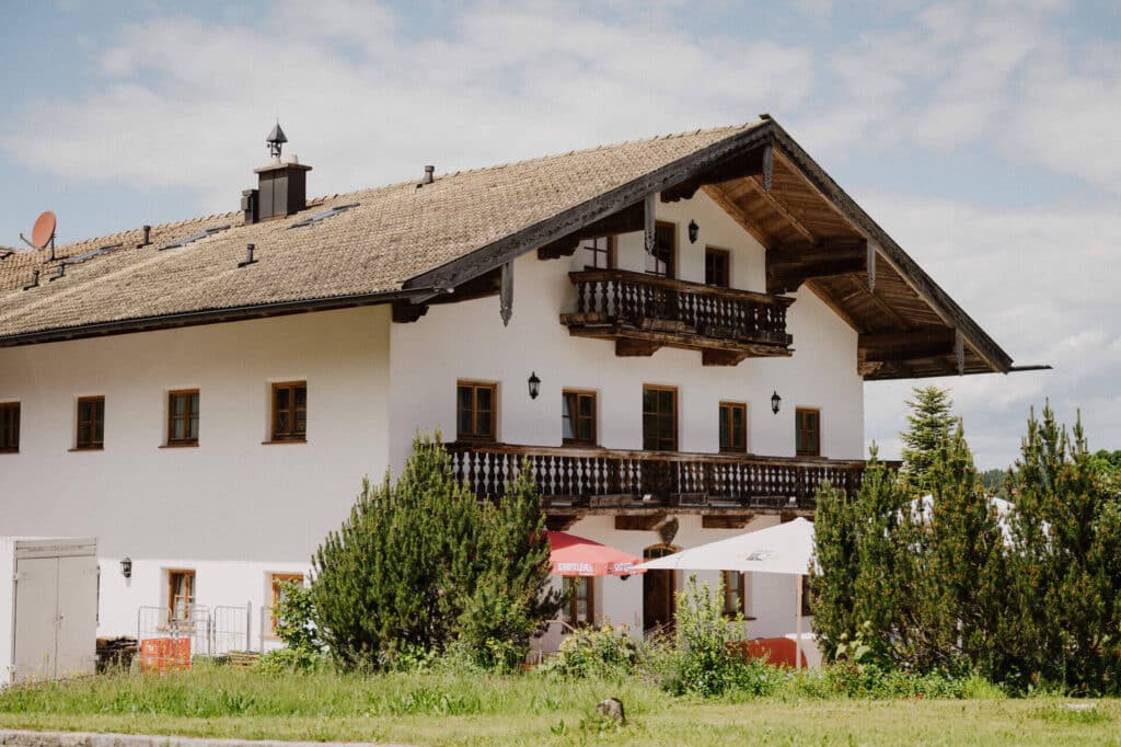 Ein großes, traditionelles Alpenhaus mit Holzbalkons und steilem Satteldach ist von Grün umgeben und perfekt für eine romantische Hochzeit. Es hat mehrere Fenster und Sonnenschirme, die einen charmanten Sitzbereich darunter vermuten lassen. Der Himmel ist teilweise bewölkt.