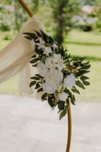 Ein dekorativer Bogen, geschmückt mit weißen Rosen, Grünpflanzen und goldenen Akzenten vor einer üppigen, grünen Außenkulisse. Perfekt für eine Hochzeit, transparenter Stoff überzieht den runden Rahmen und schafft eine elegante und romantische Szene, ideal für Paare, die heiraten möchten.