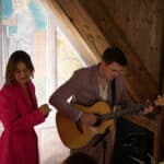 Eine Frau in einem hellrosa Mantel steht neben einem Mann von Greifbar.Eventband, der eine akustische Gitarre spielt. Sie befinden sich in einem Innenraum, in der Nähe eines Holzbalkens und einer Wand mit gemalten religiösen Figuren. Warmes Licht füllt den gemütlichen, rustikalen Raum.