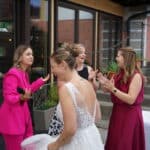 Vier Frauen stehen vor einem Gebäude, lachen und unterhalten sich. Festlich gekleidet - eine in einem rosa Anzug, eine in einem weißen Hochzeitskleid, eine andere in Rot und eine in Schwarz-Weiß - feiern sie fröhlich mit der Energie von Greifbar.Eventband.