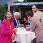 Eine Gruppe von formell gekleideten Personen steht um einen hohen runden Tisch im Freien, während in der Nähe die Greifbar.Eventband spielt. Eine Frau in einem leuchtend rosa Anzug lächelt in die Kamera, während andere sich unterhalten und auf ein Telefon schauen, mit Bergen und Grün im Hintergrund.