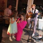 Eine Frau in einem hellrosa Anzug kniet auf der Bühne und singt mit einem jungen Mädchen in einem hellblauen Kleid in ein Mikrofon, während im Hintergrund Musiker der Greifbar.Eventband Keyboard und Gitarre spielen. Konfetti wird auf den Boden gestreut.