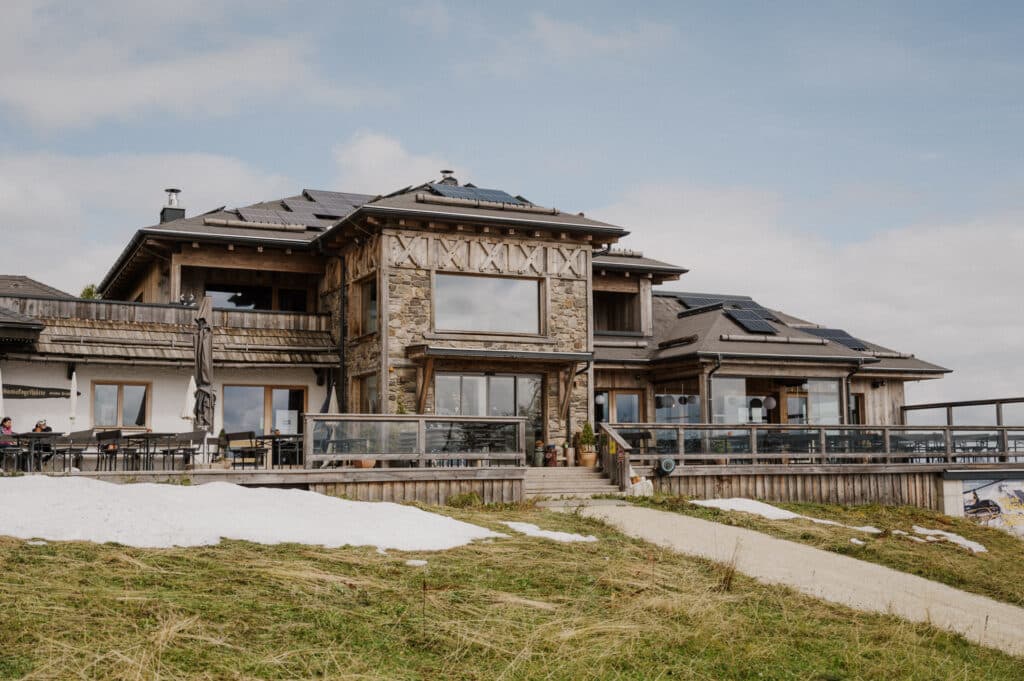 Ein großes, rustikales Gebäude im Chalet-Stil mit Solarmodulen auf dem Dach liegt auf einem grasbewachsenen Hügel mit Schneeflecken, perfekt für eine romantische Hochzeit. Auf der linken Seite lockt eine Terrasse mit Tischen und Stühlen unter einem teilweise bewölkten Himmel.