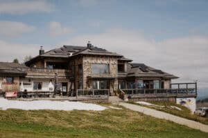 Ein rustikales Bergchalet mit einer Außenfassade aus Holz und Stein, perfekt für eine Traumhochzeit. Es verfügt über große Fenster und Sonnenkollektoren auf dem Dach, umgeben von einer hügeligen Landschaft mit Schnee- und Grasflecken unter einem teilweise bewölkten Himmel, ideal für eine durchdachte Hochzeitsplanung.