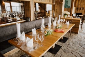 Ein gemütliches Restaurant mit einem langen Holztisch, der für ein Hochzeitsessen gedeckt ist. Er ist mit gefalteten weißen Servietten, Gläsern und einem Blumenarrangement ausgestattet. Auf dem Tisch steht eine rote Pizzaschachtel, perfekt für eine zwanglose Hochzeitsplanung inmitten der charmanten Holzwände und des Steinbodenambientes.