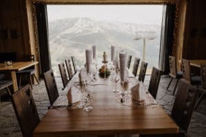 Ein für ein formelles Essen gedeckter Esstisch steht vor einem großen Fenster mit Blick auf die schneebedeckten Berge – perfekt für alle, die eine Hochzeit planen. Der Tisch ist mit weißen Servietten, Weingläsern, Menükarten und einem Blumenarrangement in der Mitte geschmückt. Gemütliche Holzstühle umgeben den Tisch.