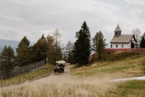 Eine Pferdekutsche fährt einen Feldweg entlang zu einer malerischen Kapelle, die sich perfekt für eine intime Hochzeit eignet, umgeben von Bäumen und Hügeln. Der bewölkte Himmel trägt zum ruhigen und rustikalen Charme dieser idyllischen Umgebung bei, die ideal für eine romantische Hochzeitsplanung im Herzen der Natur ist.
