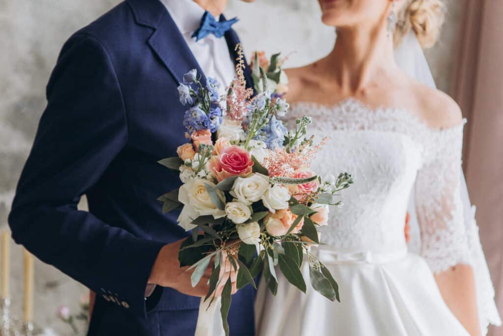 Braut und Bräutigam stehen eng beieinander und halten einen Strauß rosa, weißer und blauer Blumen in den Händen. Die Braut trägt ein schulterfreies Spitzenhochzeitskleid, während der Bräutigam einen marineblauen Anzug mit blauer Fliege trägt. „Willkommen“ wird geflüstert, während sich der Moment vor dem leicht verschwommenen Hintergrund entfaltet.