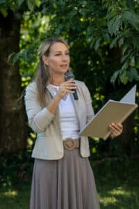 Eine Frau steht im Freien, hält in der einen Hand ein Mikrofon und in der anderen einen offenen Ordner und fängt bezaubernde Momente ein. Sie trägt einen leichten Blazer und einen langen Rock. Bäume im Hintergrund suggerieren eine natürliche Kulisse und verstärken die bezaubernde Atmosphäre.