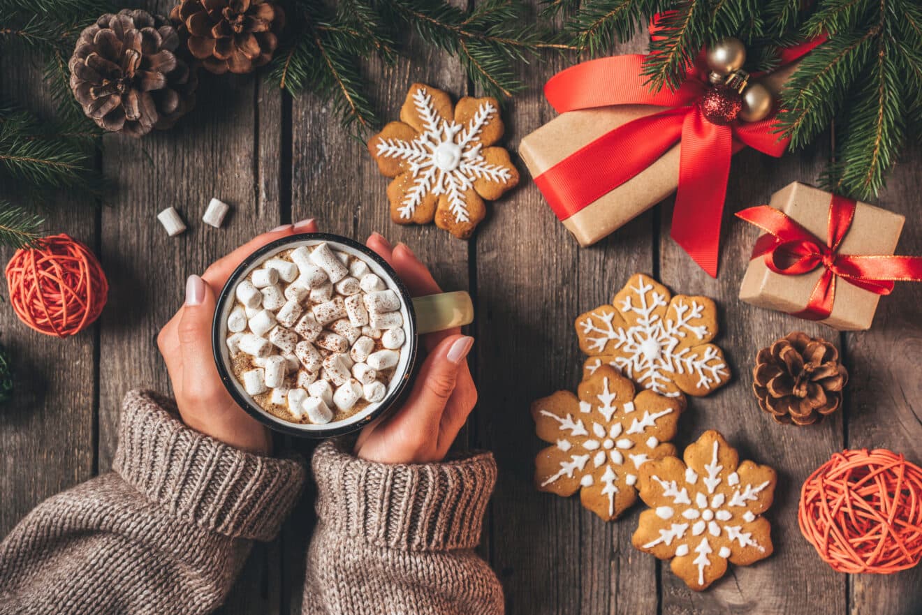 Hände halten eine Tasse heiße Schokolade mit Marshmallows. Umgeben von Lebkuchen, Tannenzapfen, verpackten Geschenken mit roten Bändern und Tannenzweigen auf einem Holztisch. Gemütliche, festliche Atmosphäre.