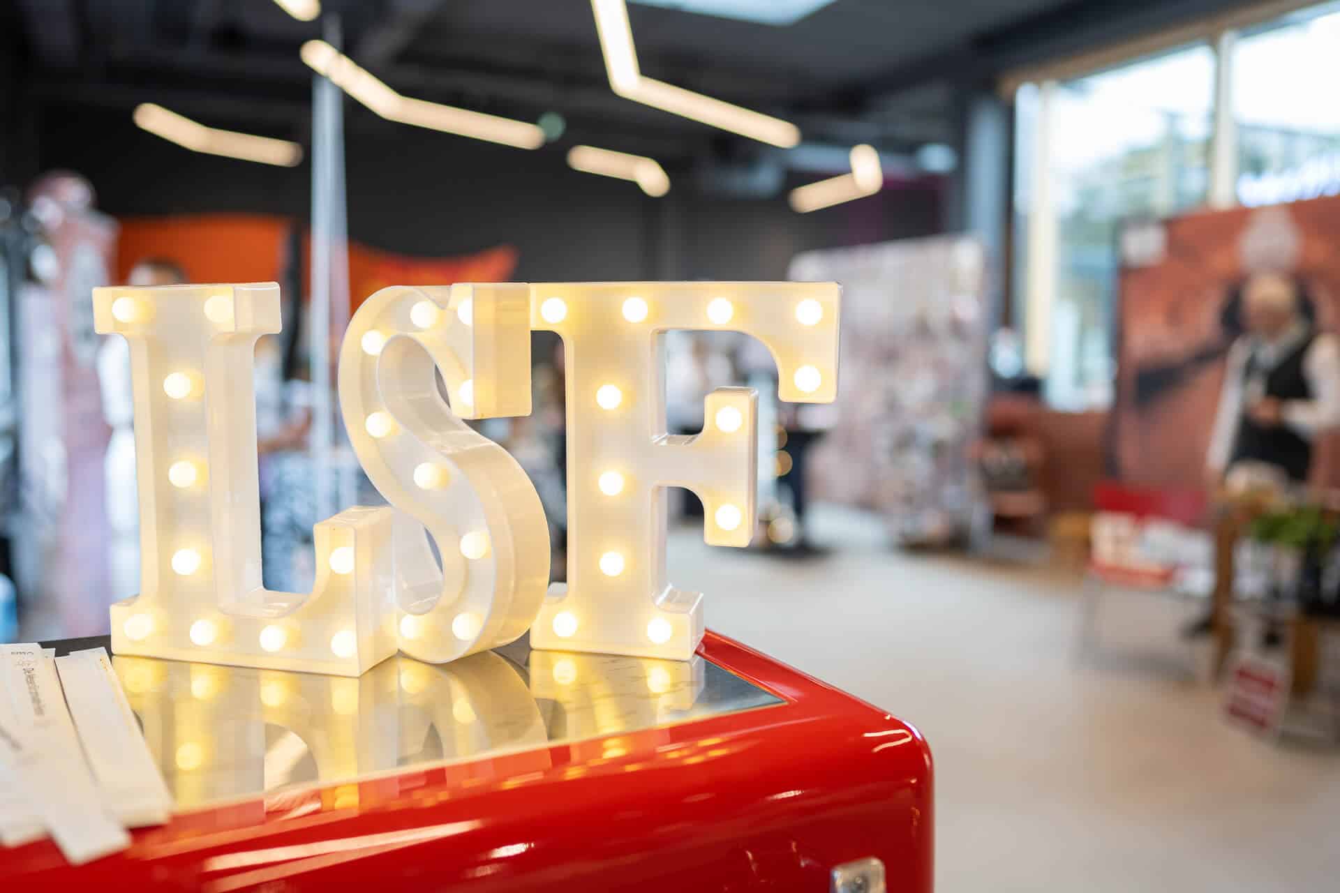 Die beleuchteten Buchstaben „LSF“ stehen auf einer glänzenden roten Oberfläche in einem modernen, geräumigen Raum. Der Hintergrund ist verschwommen, verschiedene Gegenstände und Personen sind leicht zu erkennen, was eine zwanglose oder ereignisreiche Atmosphäre suggeriert.