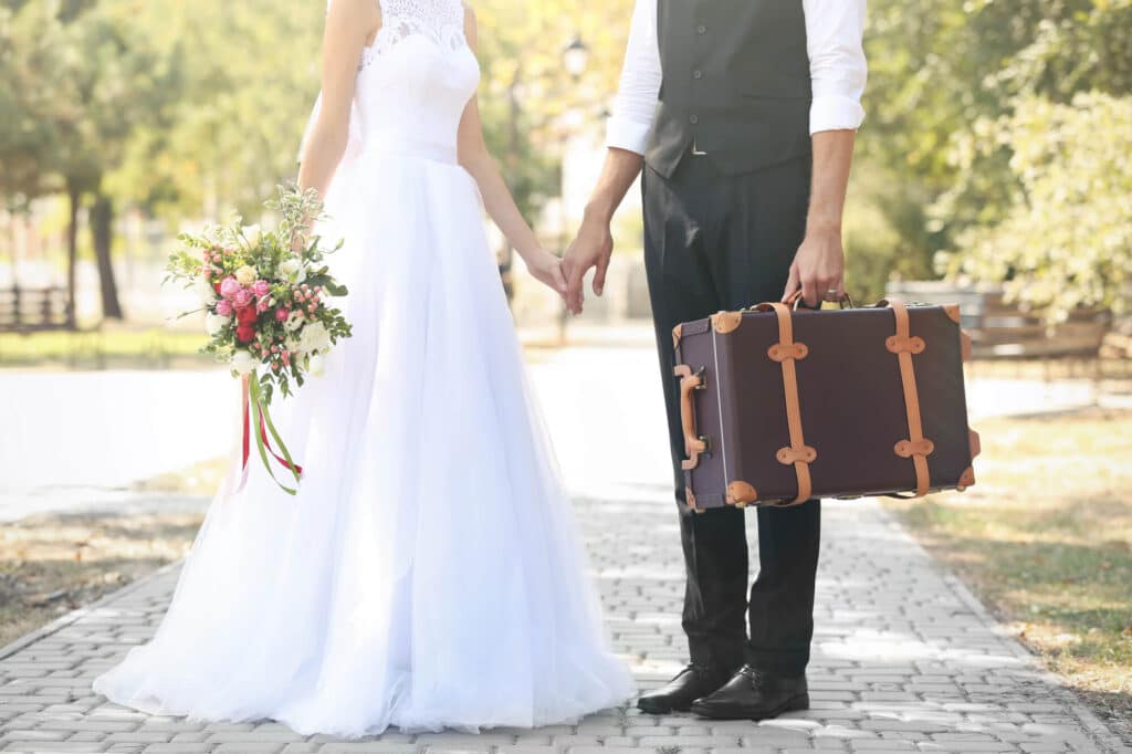 Eine Braut im weißen Kleid und ein Bräutigam in Weste und Hose stehen Händchen haltend auf einem sonnigen Weg und sind bereit, ihre Flitterwochen zu beginnen. Die Braut hält einen bunten Blumenstrauß in der Hand, der Bräutigam trägt einen Vintage-Koffer. Im Hintergrund säumen Bäume den Weg.