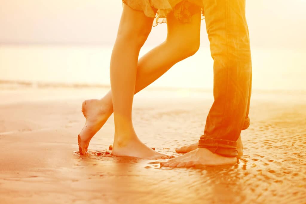 Ein Paar umarmt sich während seiner Flitterwochen an einem sonnigen Strand. Das Bild konzentriert sich auf ihre Beine und Füße, wobei die Frau auf Zehenspitzen steht. Sand und Wasser sind sichtbar und erzeugen durch das Sonnenlicht einen warmen, goldenen Schimmer.