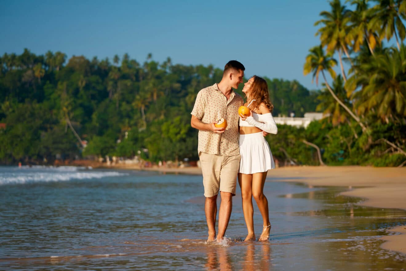 Ein Paar auf Hochzeitsreise schlendert barfuß an einem tropischen Strand entlang und lächelt mit Getränken in ausgehöhlten Früchten. Der Mann trägt ein geblümtes Hemd und Shorts, während die Frau in einem weißen schulterfreien Kleid glänzt. Im Hintergrund wiegen sich Palmen unter einem strahlend blauen Himmel.
