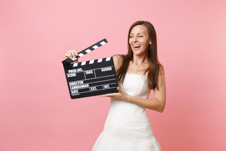 Eine Frau in einem weißen Brautkleid lächelt und hält eine Filmklappe vor einem rosa Hintergrund. Sie sieht aufgeregt und fröhlich aus – bereit, das perfekte Hochzeitsvideo zu erstellen.
