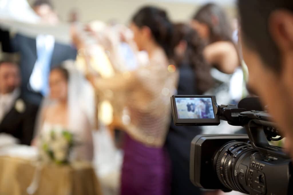 Eine Nahaufnahme einer Videokamera, die eine formelle Veranstaltung aufzeichnet – vielleicht ein Hochzeitsvideo – mit elegant gekleideten Gästen im Hintergrund, darunter eine Frau in einem goldenen Oberteil und einem lila Rock; der Kamerabildschirm zeigt die aufgezeichnete Szene.