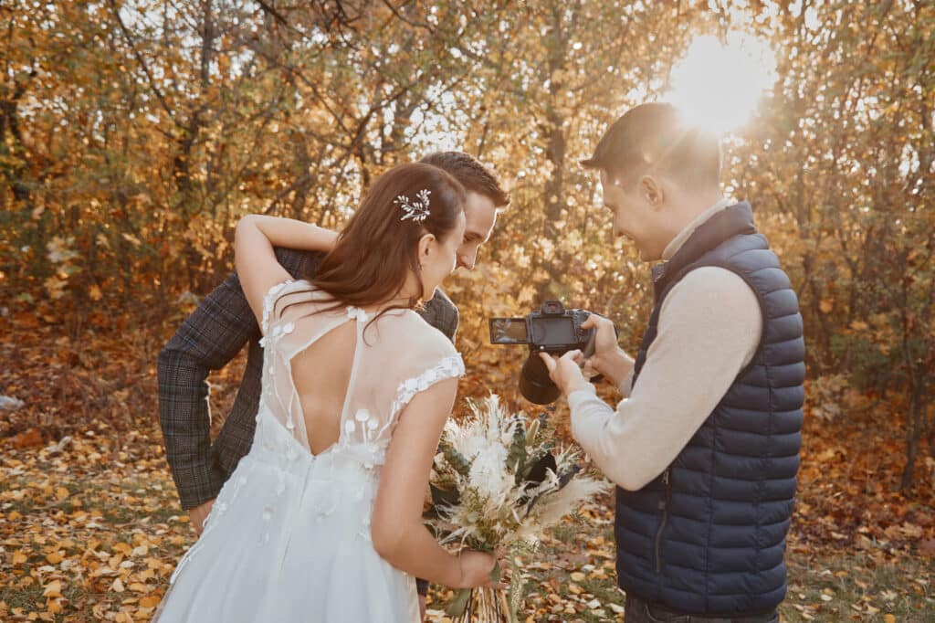 Braut und Bräutigam im Hochzeitskleid schauen sich ihr Hochzeitsvideo im Freien mit einem Fotografen auf dem Kamerabildschirm an. Herbstlaub und Sonnenlicht füllen den Hintergrund und schaffen eine warme, goldene Atmosphäre.