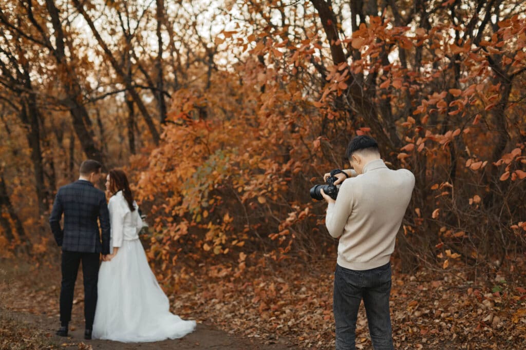Ein Fotograf filmt Braut und Bräutigam, wie sie Händchen halten und in einem Wald, der im warmen Herbstlaub leuchtet, den Blick abwenden – die perfekte Inspiration für ein romantisches Hochzeitsvideo.